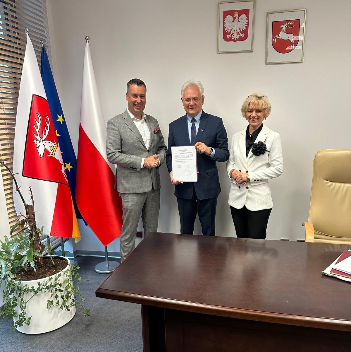 Na zdjęciu widzimy trzy osoby stojące w eleganckim biurze na tle flag i godła.
W centrum kadru stoi mężczyzna w ciemnym garniturze, trzymający w rękach dokument skierowany przodem do kamery – to prawdopodobnie Uchwała Zarządu Województwa. Z jego wyrazu twarzy można wyczytać powagę i satysfakcję.
Po jego lewej stronie (z prawej strony patrzącego) stoi kobieta w białym garniturze z czarnym kwiatem przypiętym do klapy marynarki. Uśmiecha się serdecznie w stronę obiektywu.

Po prawej stronie mężczyzny (z lewej strony patrzącego) stoi inny mężczyzna – w szarym garniturze, z ciemnymi włosami i również uśmiechnięty. Lekko dotyka ramienia osoby w centrum i gestykuluje dłonią, jakby właśnie złożył gratulacje.
W tle widoczne są:
– trzy flagi (flaga województwa lubelskiego, flaga Polski i flaga Unii Europejskiej),
– dwa godła na ścianie: orzeł biały (godło Polski) i herb województwa lubelskiego – biały jeleń na czerwonym tle.
Po lewej stronie kadru widoczna jest roślina doniczkowa, a po prawej duże biurko z dokumentami.
Całość utrzymana jest w oficjalnym, reprezentacyjnym tonie – to wyraźnie moment wręczenia ważnego dokumentu w instytucji publicznej.