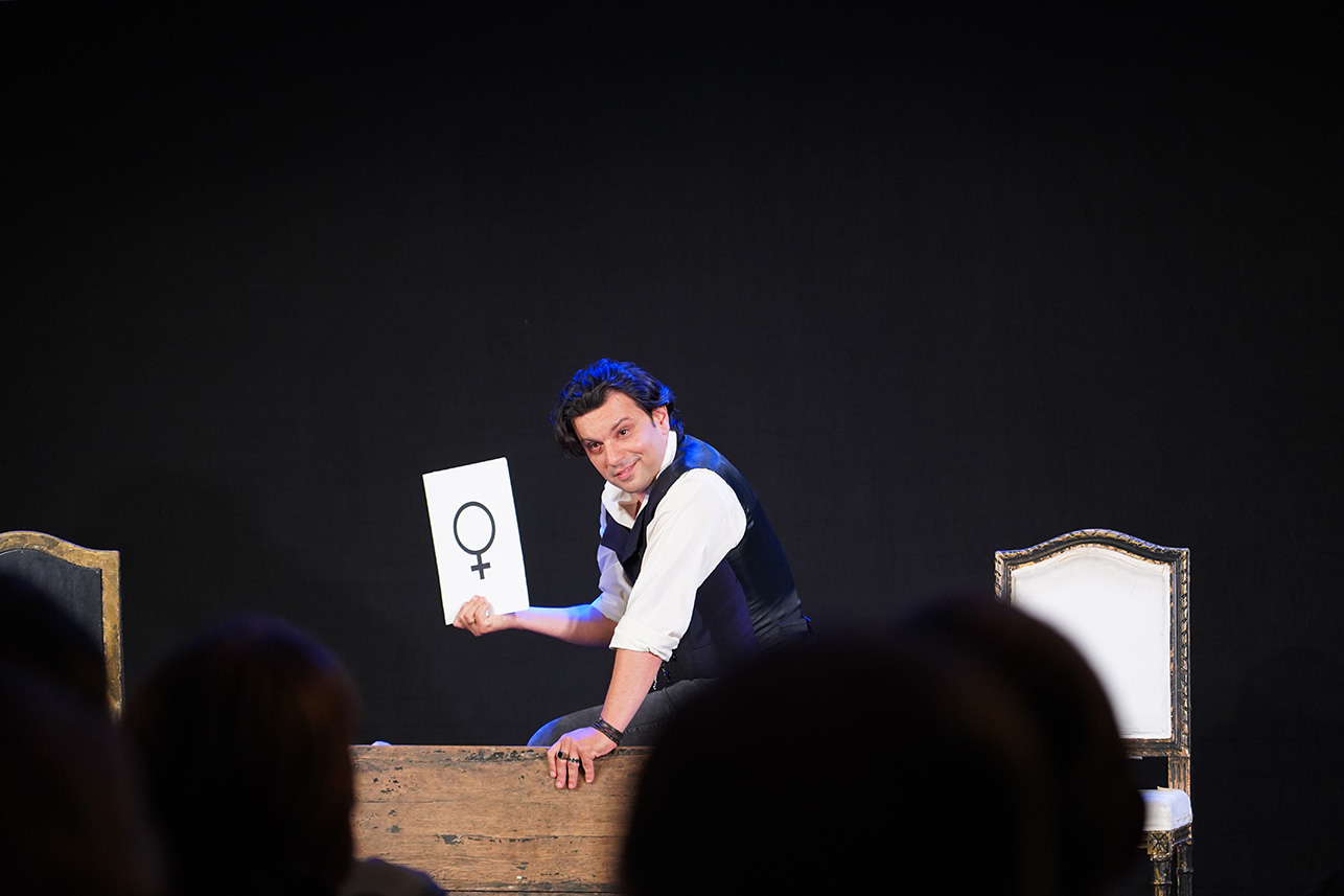 Podsumowanie spektaklu „Sonata Kreutzerowska” w Muzeum Zamoyskich w Kozłówce” Na zdjęciu widać scenę teatralną z ciemnym, jednolitym tłem. Na pierwszym planie znajduje się aktor – mężczyzna w średnim wieku, o ciemnych włosach, ubrany w białą koszulę z podwiniętymi rękawami i ciemną kamizelkę. Aktor siedzi bokiem na drewnianej skrzyni lub niskim podwyższeniu i patrzy bezpośrednio w stronę publiczności z lekkim, sugestywnym uśmiechem.
W jego prawej dłoni uniesionej w górę znajduje się biała kartka lub plansza z dużym, czarnym symbolem kobiety (okrąg z krzyżykiem poniżej). Symbol ten jest wyraźnie widoczny i stanowi główny element przekazu wizualnego.
W tle, po obu stronach aktora, stoją dwa stylizowane krzesła o klasycznym wyglądzie – jasne siedziska i oparcia z ciemnymi, ozdobnymi ramami. Przed sceną widać zarysy głów widzów siedzących na widowni, co sugeruje, że zdjęcie zostało wykonane podczas spektaklu.
Atmosfera sceny jest intymna, skupiona, z wyraźnym teatralnym napięciem. Oświetlenie podkreśla twarz i sylwetkę aktora, a tło pozostaje w cieniu, co kieruje całą uwagę na przekaz postaci i trzymany przez nią znak.
