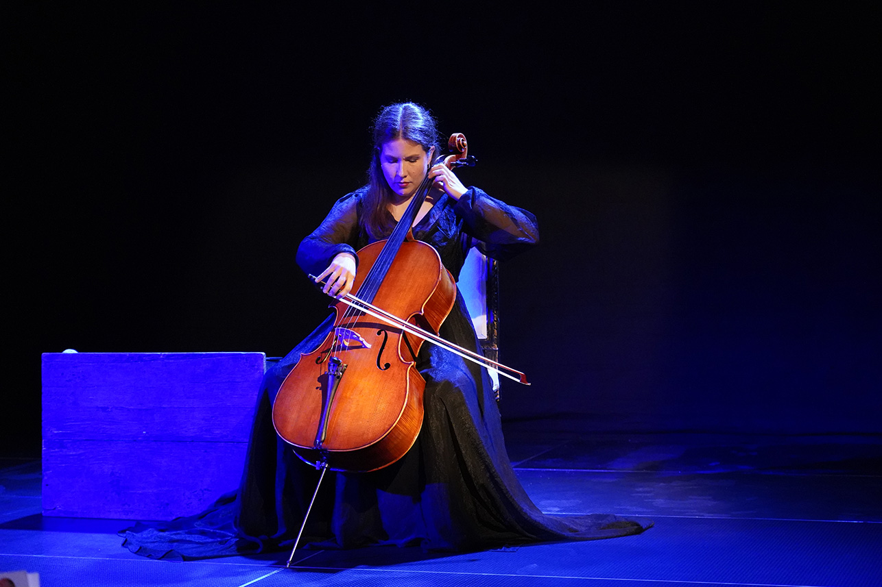 Podsumowanie spektaklu „Sonata Kreutzerowska” w Muzeum Zamoyskich w Kozłówce” Na zdjęciu widzimy młodą kobietę grającą na wiolonczeli na scenie teatralnej. Siedzi na klasycznym krześle z jasnym oparciem, ubrana w długą, czarną suknię z prześwitującymi rękawami. Jej długie, brązowe włosy są luźno rozpuszczone. Kobieta jest skupiona – pochylona lekko do przodu, z wyrazem głębokiej koncentracji na twarzy.
Wiolonczela, o klasycznym drewnianym wykończeniu, dominuje centralną część zdjęcia. Artystka trzyma smyczek w prawej dłoni i prowadzi go po strunach, lewą ręką przyciskając je na gryfie. Na scenie panuje nastrojowe, ciemne oświetlenie z chłodnym, niebieskawym światłem skierowanym na postać muzyczki i jej instrument. Tło jest jednolicie czarne, co potęguje atmosferę skupienia i emocjonalnej głębi.
Po lewej stronie kadru widoczna jest prosta drewniana skrzynia pomalowana na niebiesko, która może być elementem scenografii. Całość tworzy intymny, artystyczny klimat – scena wydaje się być częścią spektaklu teatralnego z muzyką wykonywaną na żywo.