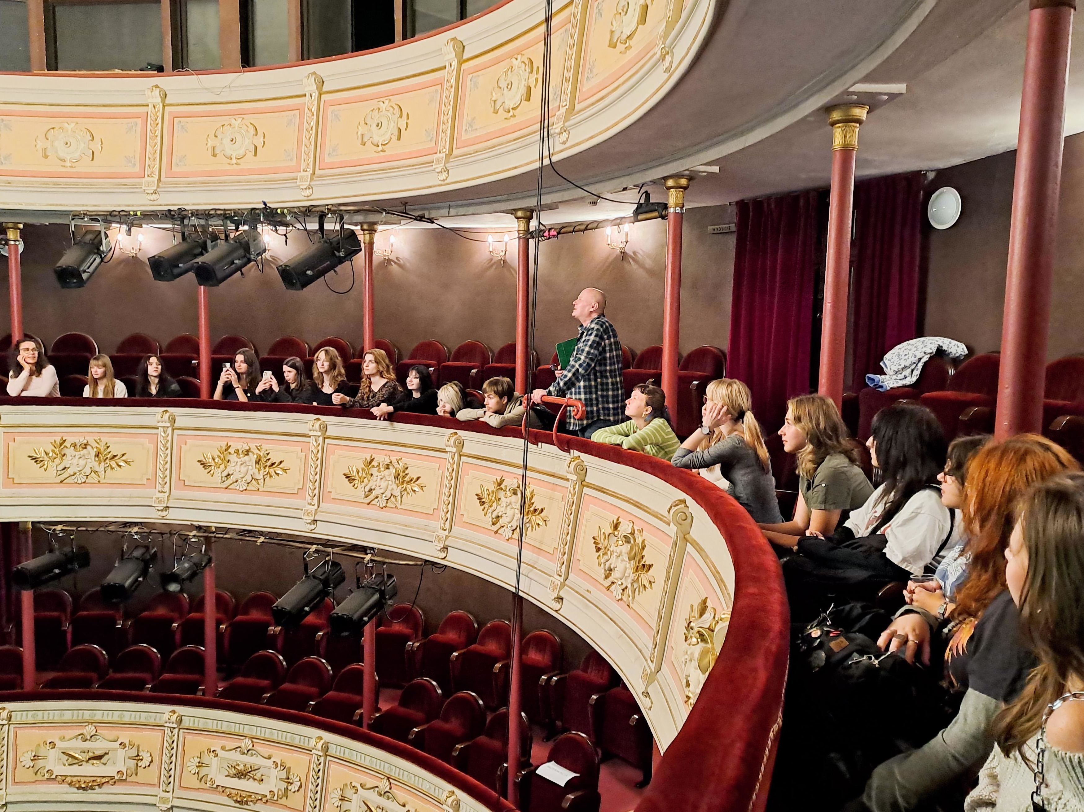 Fot. 1 Na zdjęciu widać grupę młodzieży wraz z dorosłym opiekunem, pozujących wspólnie na tle teatralnej scenografii.
Osoby siedzą i stoją na schodach ustawionych przed sceną. Grupa liczy około dwudziestu osób – większość to nastolatki, głównie dziewczęta, a wśród nich jeden starszy mężczyzna w koszuli w kratę. Wszyscy patrzą w stronę obiektywu, część uśmiecha się, inni mają poważniejsze miny.
W tle widoczna jest teatralna scenografia – dwa duże, malowane elementy przypominające fragmenty antycznej architektury z łukami. Widać na nich pejzaże: niebo, wodę, drzewa i budynki. Nad sceną rozciągnięto jasną siatkę lub materiał przypominający baldachim.
Cała grupa sprawia wrażenie zadowolonej, jakby zdjęcie było pamiątką po wspólnym wydarzeniu – najpewniej wizycie w teatrze lub warsztatach scenicznych.