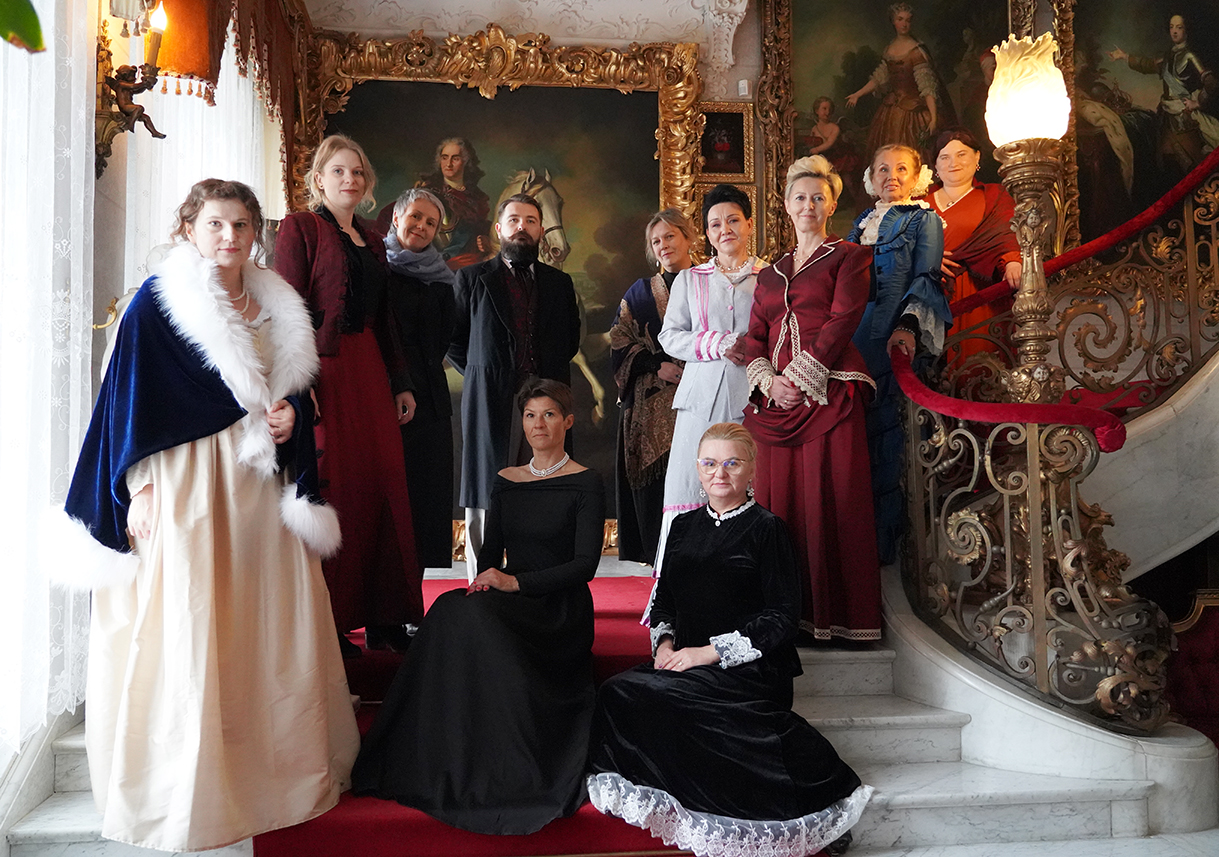 Zdjęcie przedstawia grupę dwunastu osób ubranych w eleganckie stroje historyczne, nawiązujące do XIX-wiecznej mody europejskiej. Scena rozgrywa się wewnątrz bogato zdobionego pałacu. Wszyscy stoją lub siedzą na szerokich, marmurowych schodach pokrytych czerwonym dywanem.
Po lewej stronie kadru znajduje się duże, jasne okno z koronkowymi firanami, wpuszczające do wnętrza naturalne światło. Po prawej widać kunsztowną, złotą balustradę schodów, ozdobioną rzeźbieniami i zakończoną lampą przypominającą płomień. Ściany zdobią monumentalne portrety w pozłacanych ramach, przedstawiające możnowładców.
W pierwszym planie dwie kobiety siedzą na schodach. Po lewej — w długiej, czarnej sukni z odkrytymi ramionami i krótką perłową kolią przy szyi; po prawej — w czarnej, aksamitnej sukni z białymi koronkami przy rękawach i dole spódnicy, z jasnymi włosami upiętymi i okularami.
Za nimi, na wyższych stopniach, stoi grupa dziesięciu osób. Po lewej — kobieta w kremowej sukni i granatowym płaszczu z białym futrzanym wykończeniem. Obok niej — kobieta w bordowej sukni z czarnym gorsetem. W centrum stoi mężczyzna z brodą w czarnym surducie. Wokół niego — kolejne kobiety w długich, zdobionych sukniach: bordowych, granatowych, szarych i niebieskich. Stroje są zróżnicowane, pełne detali — koronek, guzików i ozdobnych kołnierzy.
Wszyscy patrzą bezpośrednio w obiektyw, prezentując się dostojnie i poważnie, jak do oficjalnego portretu. Atmosfera fotografii przywołuje skojarzenia z balem lub wystawnym spotkaniem arystokracji.