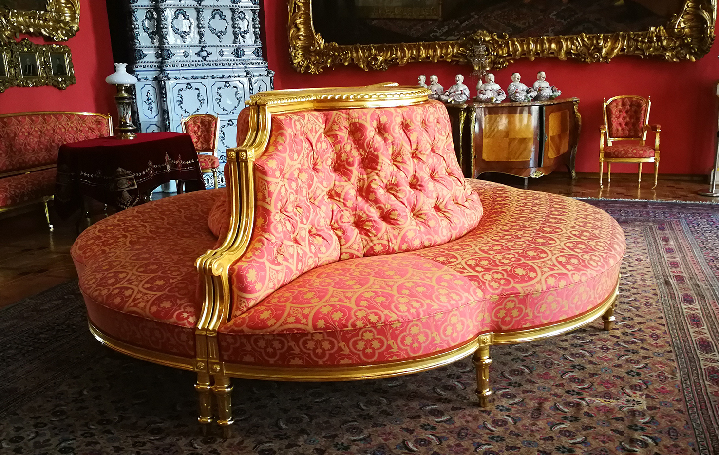 Dane katalogowe obiektu
Kanapy półokrągłe w stylu Ludwika XVI
Sygnowane przez Magazyn mebli i gotowych robót tapicerskich Leopolda Mergentalera
Datowanie druga połowa dziewiętnastego wieku
Tworzywo drewno, tkanina żakardowa
Technika złocenie, tapicerowanie, snycerka
Opis mebli
Na fotografii widoczne są dwie stylowe kanapy o półokrągłym kształcie, zestawione ze sobą w taki sposób, że tworzą jeden okrągły mebel. Mają niską, szeroką formę. Oparcie jest wysokie, miękkie i pikowane – na całej powierzchni widać regularnie rozmieszczone guziki tworzące charakterystyczne zagłębienia i wypukłości, co daje puszystą, niemal poduszkową strukturę. Siedzisko jest obszerne, dopasowane do zaokrąglonej formy całej konstrukcji. Górna krawędź oparcia oraz boczne wykończenia wykonane są z rzeźbionego drewna w kolorze złotym. Kanapy stoją na toczonych, smukłych nóżkach również w złotym kolorze.  Tapicerka ma intensywny, ciepły odcień czerwieni, pokryty regularnym, złotym wzorem roślinnym. Wzór tworzą stylizowane liście winorośli i czterolistne motywy zamknięte w powtarzalnych, symetrycznych kształtach. W tle stojących kanap widok salonu, wyposażonego w stylowe meble, bogato zdobiony piec kaflowy oraz liczne obrazy w ramach koloru złotego.