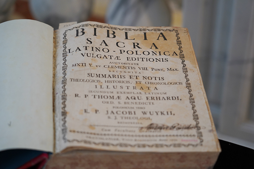 Na zdjęciu widać starą, otwartą książkę. Jest to stary egzemplarz Biblii, z pożółkłymi stronami i ozdobnym tytułem po łacinie na pierwszej stronie.