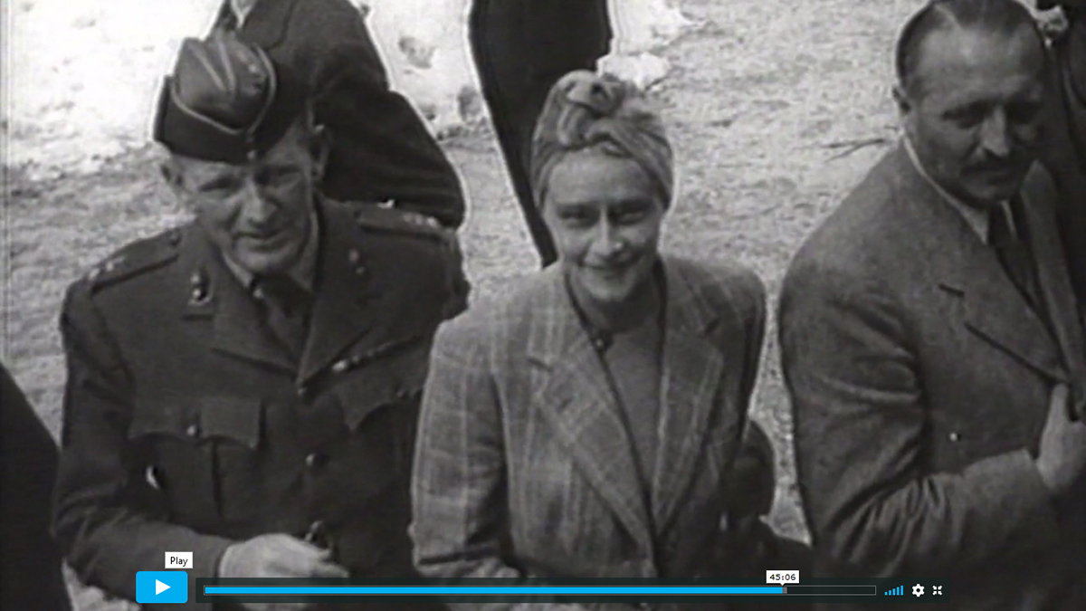 Kadr z dokumentu fabularyzowanego „Wir Geiseln der SS”, (Jesteśmy zakładnikami SS). Dwóch mężczyzn i kobieta stojąca pomiędzy nimi. Mężczyzna po lewej stronie, w średnim wieku, ubrany w mundur, na głowie furażerka. Kobieta, młoda, ubrana w jasny żakiet w kratę, na głowie modnie upięty turban. Uśmiecha się. Mężczyzna po prawej stronie, ustawiony prawym profilem z głową skierowana en face. Ubrany w jasna marynarkę, modnie zaczesany, z wąsem.