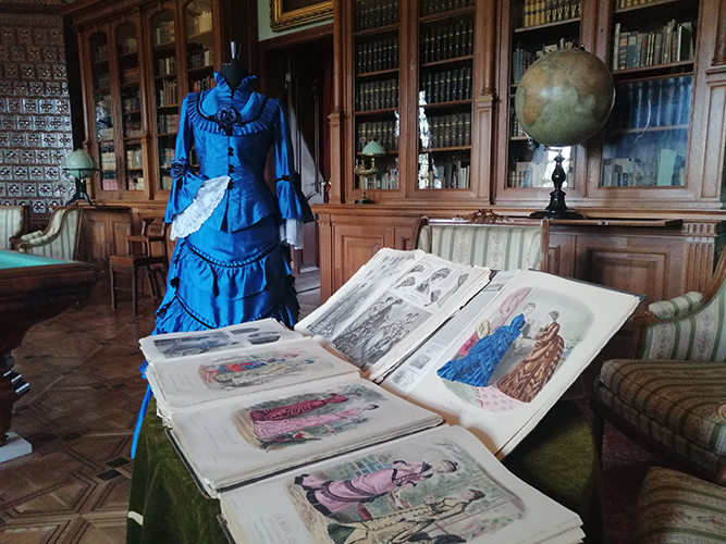 Fragment pomieszczenia z szafami bibliotecznymi. Na ich tle manekin ubrany w stylową suknię. Na pierwszym planie kilka numerów dziewiętnastowiecznego czasopisma modowego „La Mode Illustrée. Journal de la Famille”