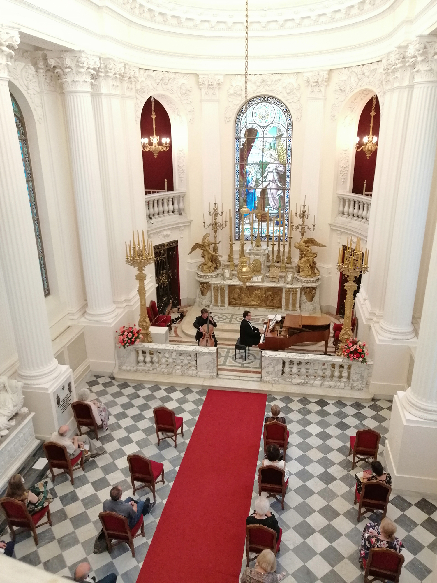 Fotografia z wysokości. Wnętrze jasnej kaplicy. W prezbiterium dwóch artystów. Po prawej stronie mężczyzna grający na fortepianie, po lewej stronie na wiolonczeli. W nawie głównej siedzący pojedynczo, w równych odległościach od siebie słuchacze.