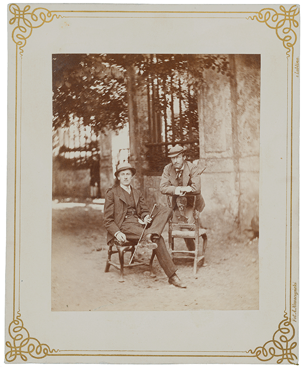 Fotografia zabytkowa, pionowa. Dwaj elegancko ubrani, młodzi mężczyźni przed bramą pałacową w Kozłówce. Mężczyzna po lewej stronie siedzi na krześle, z nogami skrzyżowanymi na wysokości kolan. W lewej ręce trzyma laseczkę. Mężczyzna po prawej ciężar ciała składa na oparcie krzesła podpierając się o nie ramionami.