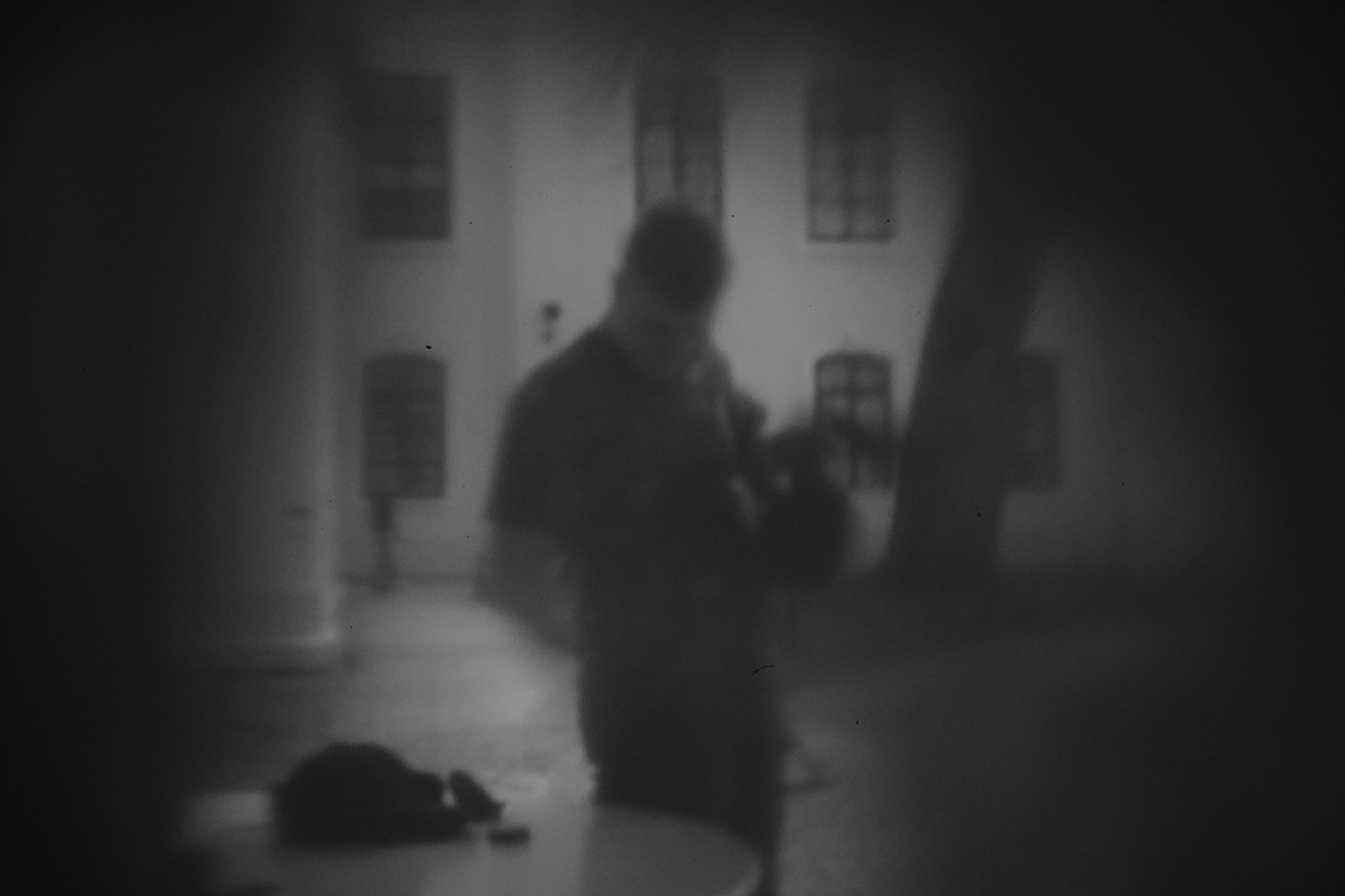 Postać młodego mężczyzny stojącego przy stoliku. W tle fragment drzewa i budynku. Fotografia wykonana techniką camera obscura, czarno-biała, lekko rozmyta, zaciemniona na rogach.