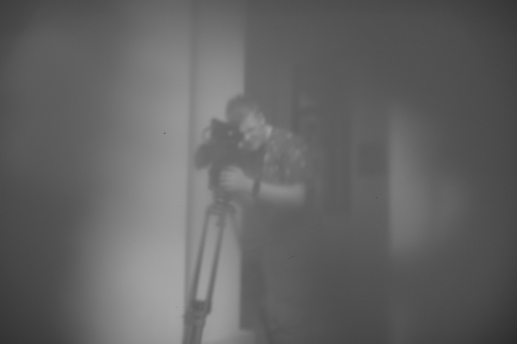 Postać młodego mężczyzny patrzącego przez ustawioną na statywie kamerę. W tle ściana budynku z oknem. Fotografia wykonana techniką camera obscura, czarno-biała, lekko rozmyta, zaciemniona na rogach.