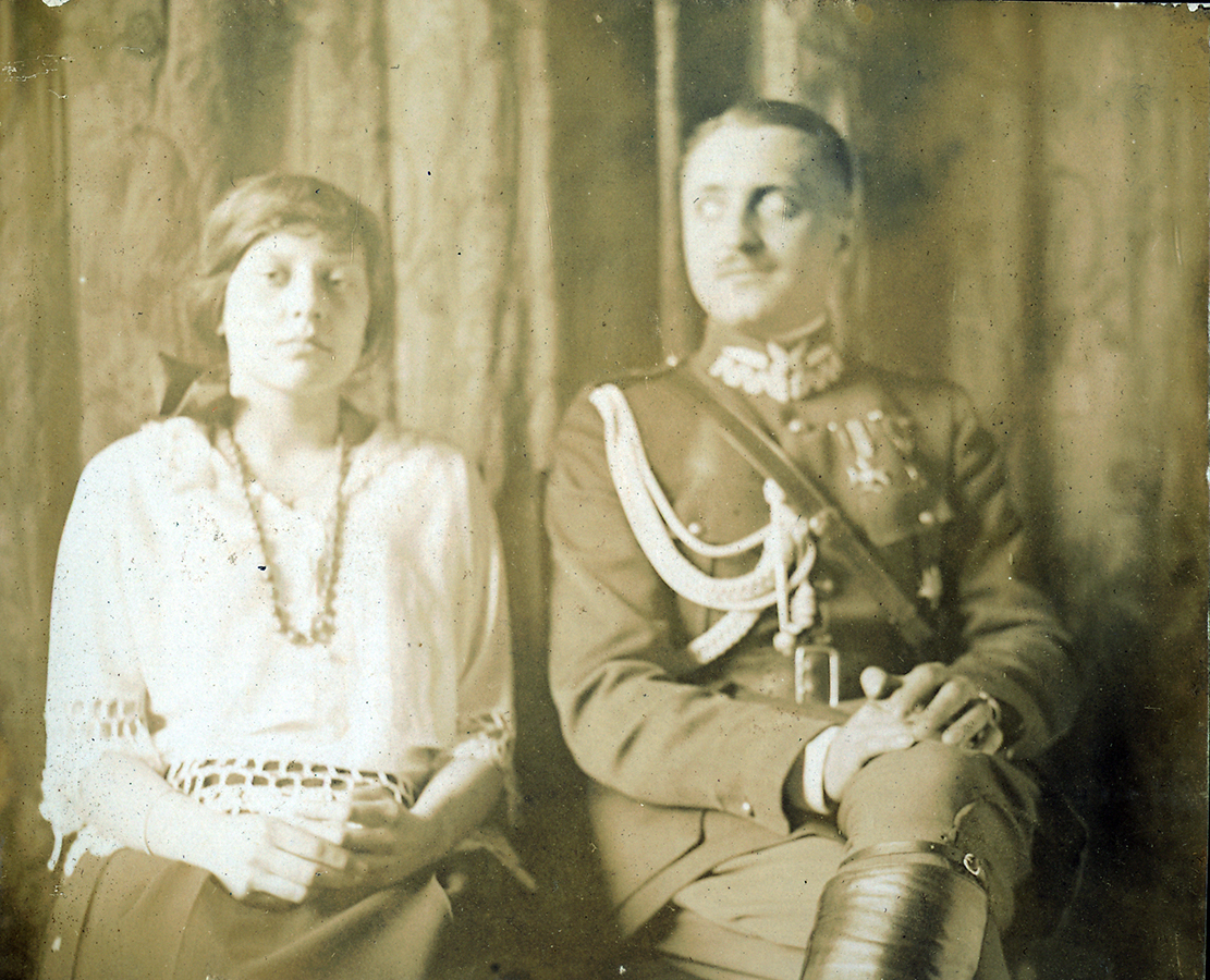 Fotografia archiwalna przedstawiająca Jadwigę i Aleksandra Leszka Zamoyskich siedzących obok siebie. Po lewej stronie młoda kobieta ubrana w białą bluzkę i ciemną spódnicę, wzrok skierowany na fotografa, dłonie splecione na kolanach. Po prawej stronie, młody mężczyzna, w galowym mundurze, głowa lekko skręcona w lewą i wzrok skierowany w dal. Noga założona na nogę, dłonie splecione na kolanach.