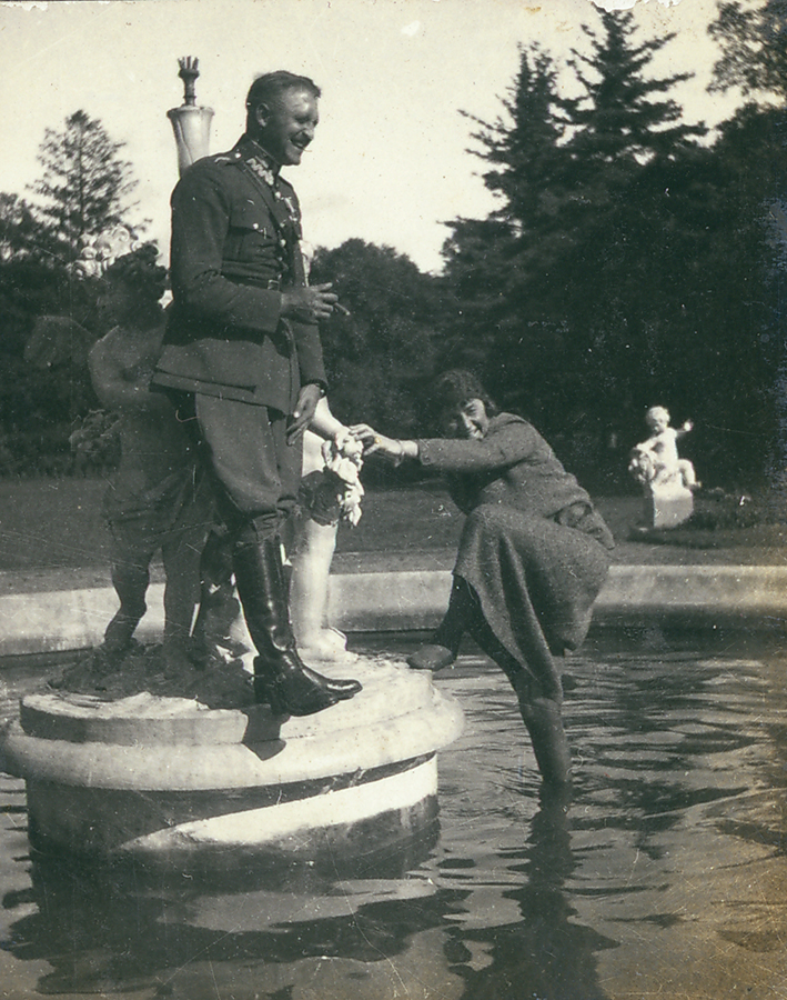 Fotografia archiwalna, czarno-biała, przedstawiająca Jadwigę i Aleksandra Leszka Zamoyskich przy fontannie w parku w Kozłówce. Na pierwszym planie młody, szeroko uśmiechnięty mężczyzna stoi na cokole, opiera się o jedną z trzech białych rzeźb fontanny. Ubrany jest w buty z cholewami i mundur oficerski ułana krechowieckiego, bez nakrycia głowy. W prawej ręce trzyma zapalone cygaro, lewą opiera na udzie. Po jego lewej stronie młoda kobieta stoi prawą nogą w wodzie, lewą opiera o cokół, przytrzymując się lewą ręką o rzeźbę fontanny. Ubrana jest w ciemny strój i ciemne pończochy.