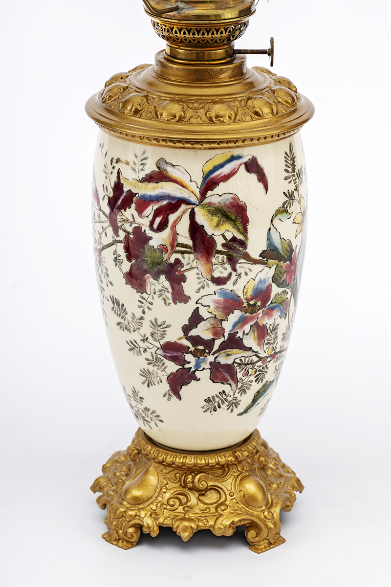 Fajansowy korpus lampy naftowej, w kształcie wydłużonej wazy, o metalowej podstawie i zbiorniku. Powierzchnia korpusu udekorowana jest kwiatami katlei w odcieniach różu, błękitu i żółci.