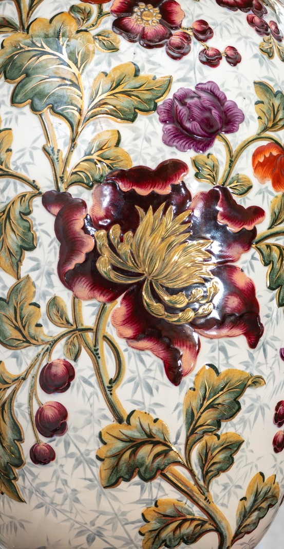 Fotografia przedstawia fragment korpusu lampy. Duży wzór kwiatowy malowany na porcelanie, z czerwonych i fioletowych kwiatów. Między nimi gałązka z okazałymi liśćmi, żyłkowanie zaznaczone złoceniami. W tle delikatnie zaznaczone szare gałązki.
