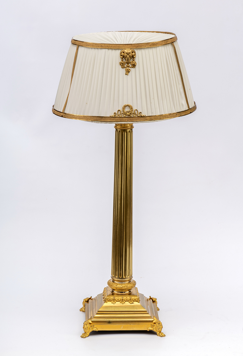 Fotografia przedstawia lampę o kolumnowej podstawie w kolorze złotym z jasnym abażurem z tkaniny rozpiętej na dekorowanych obręczach.