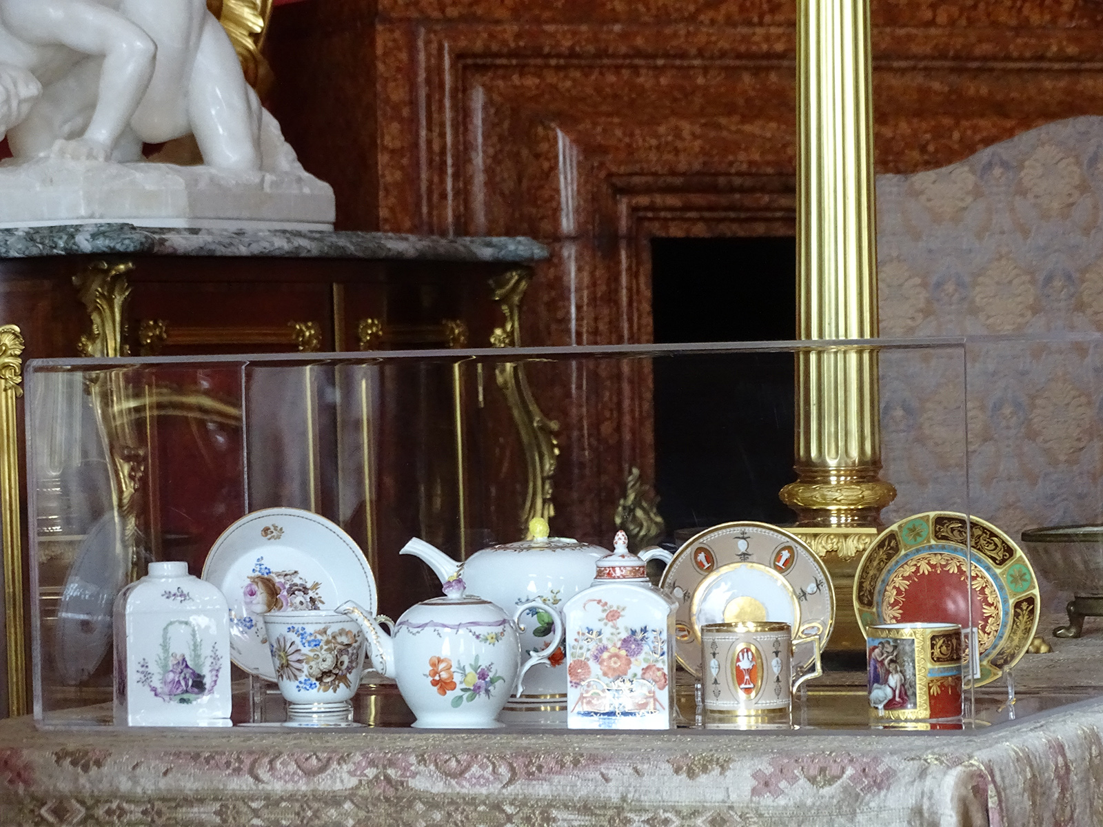 Fotografia przedstawia naczynia porcelanowe umieszczone w przezroczystym pudełku. Od lewej: biała herbatnica z kwiatową dekoracją, biała filiżanka ze spodkiem z kwiatową dekoracją, dwa białe czajniczki różnych rozmiarów z kwiatową dekoracją, biała herbatnica z dekoracją kwiatową, filiżanka ze spodkiem, w kolorze jasnobrązowym, zdobiona. Filiżanka ze spodkiem różnokolorowa (czerwień, zieleń, czerń) ze złotym ornamentem.