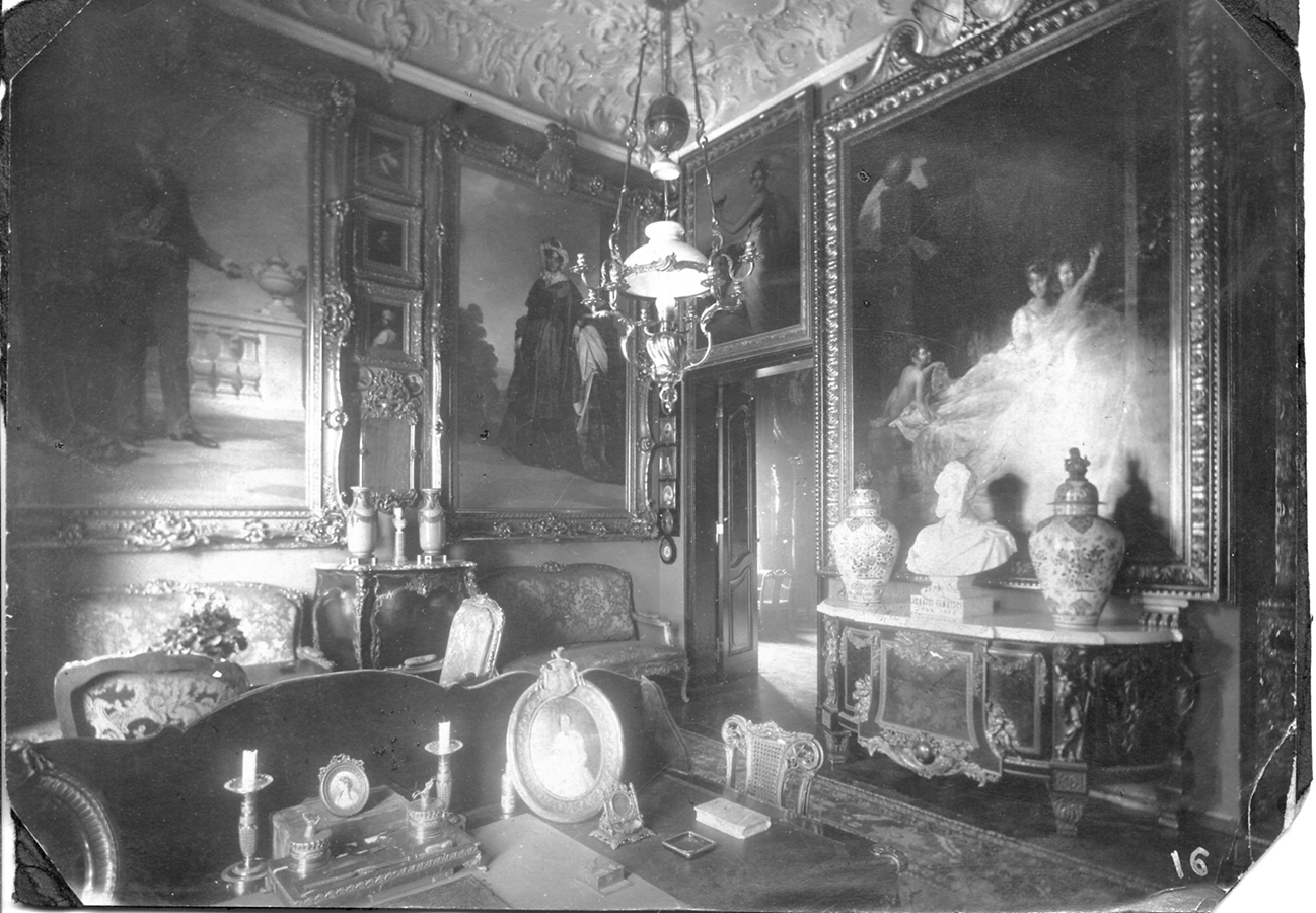Czarno-biała fotografia archiwalna przedstawiająca wnętrze Gabinetu hrabiego Konstantego Zamoyskiego około 1900 roku. Na pierwszym planie widoczny fragment biurka, na nim przybory do pisania i oprawione w stylowe ramki wizerunki matki i żony hrabiego. W głębi za biurkiem stylowe meble, po prawej stronie bogato zdobiona okazała komoda. Na niej białe marmurowe popiersie i dwie wazy. Obok widoczne otwarte drzwi prowadzące do kolejnego pomieszczenia. Na ścianach gęsto zawieszone różnej wielkości obrazy oprawione w dekoracyjne ramy. Z sufitu zwisa lampa naftowo-świecznikowa na dekoracyjnych łańcuchach.