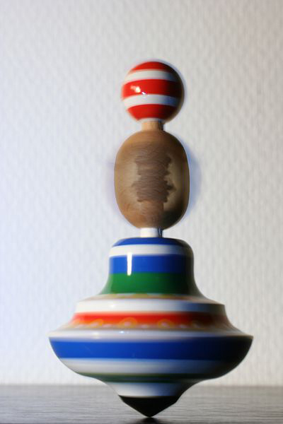 Fotografia ukazuje ceramiczną zabawkę – bąka wprawianą w ruch obrotowy dookoła swojej osi. Dolna część ma kształt spłaszczonej cebuli zakończonej szpicem, malowanej w pasy w kolorze niebieskim, zielonym i czerwonym, kolejny element podłużny w kolorze drewna, zwieńczenie stanowi kulka w czerwono białe pasy.