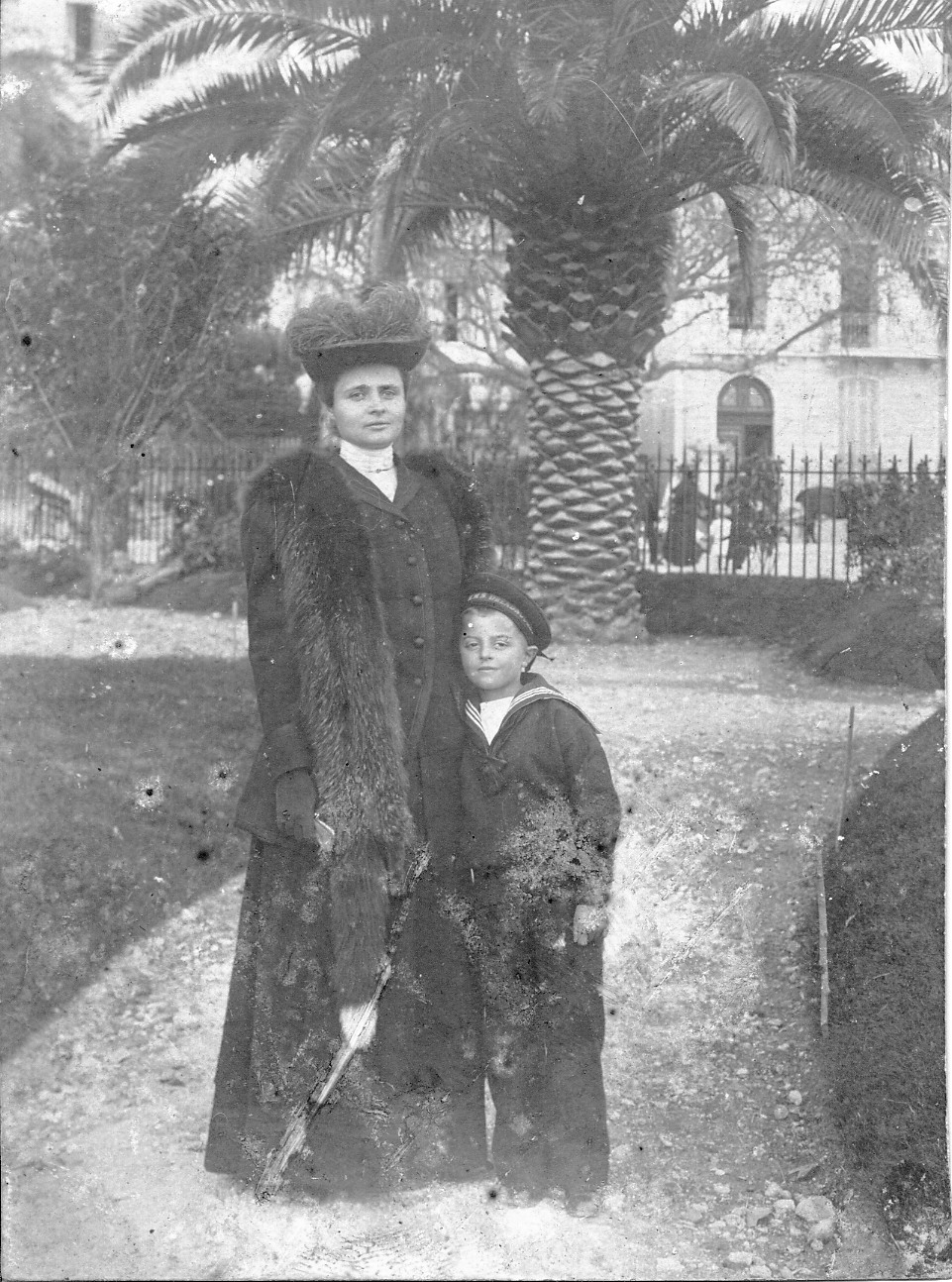 Fotografia archiwalna, czarno-biała, wykonana około 1905 roku, przedstawia Marię Potocką, późniejszą Adamową Zamoyską, z synem Michałem. Kobieta w czarnej, długiej spódnicy i żakiecie z narzuconą futrzaną etolą i bogato zdobionym kapeluszu. Przy niej, przytulony, stoi chłopiec, ubrany w ciemny, stylizowany na marynarski strój. W tle fragmenty budynków i okazała palma.