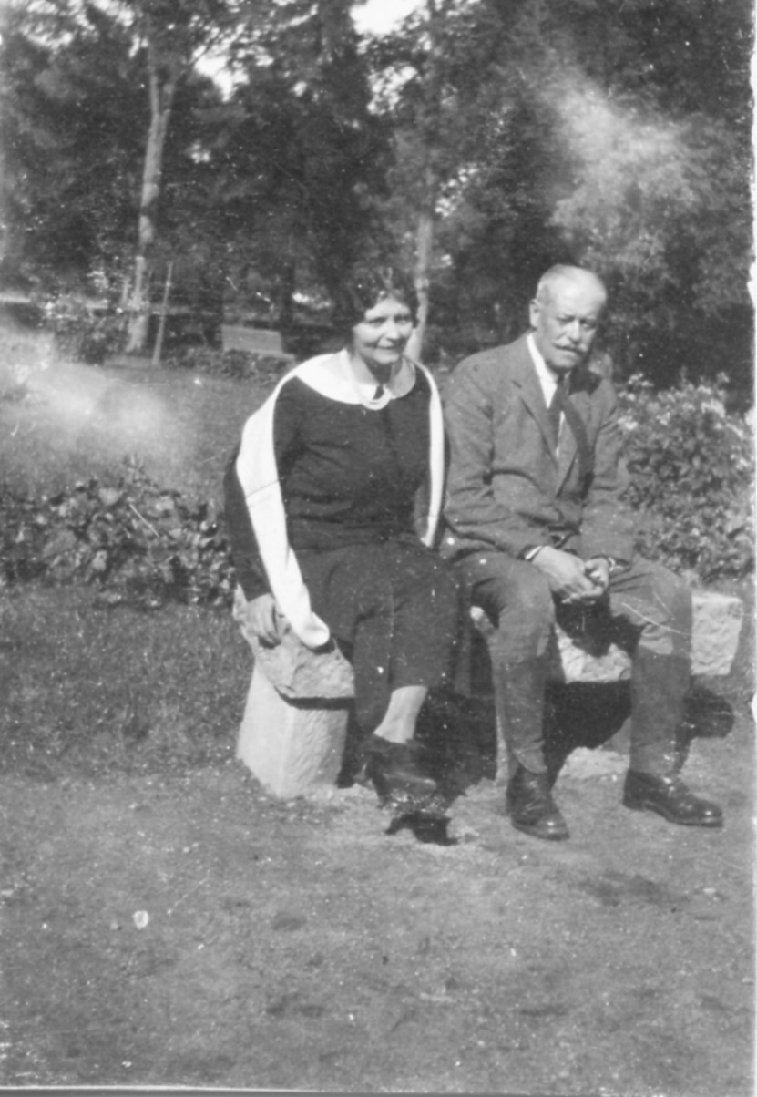 Fotografia archiwalna, czarno-biała, wykonana w latach dwudziestych XX wieku, przedstawia Marię Zamoyską i jej męża, Adama Zamoyskiego. Kobieta i mężczyzna siedzą na kamiennej ławce. Kobieta ma ciemne włosy, spięte z tyłu, ubrana jest w ciemną suknię z białym kołnierzem i biały szal. Mężczyzna ma siwe krótkie włosy i wąsy, ubrany jest w szary garnitur i jasną koszulę z krawatem.