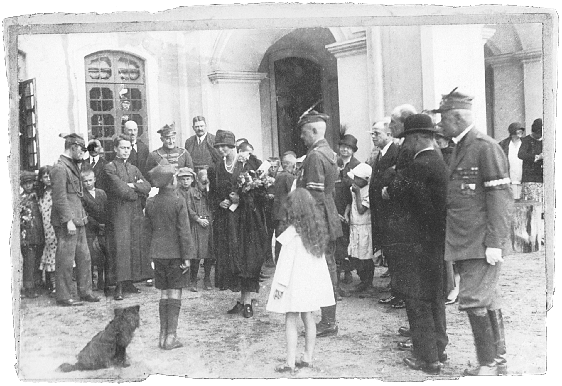 Fotografia archiwalna, czarno-biała, wykonana 2 sierpnia 1927 roku w Kozłówce, przedstawia moment wręczenia kwiatów i składania życzeń Marii Zamoyskiej z okazji imienin. Na fotografii grupa osób w różnym wieku, którzy otaczają kobietę z naręczem kwiatów i stojącego przed nią, w wyprostowanej pozie, chłopca. Niektórzy mężczyźni ubrani w mundury Towarzystwa Gimnastycznego „Sokół”.
