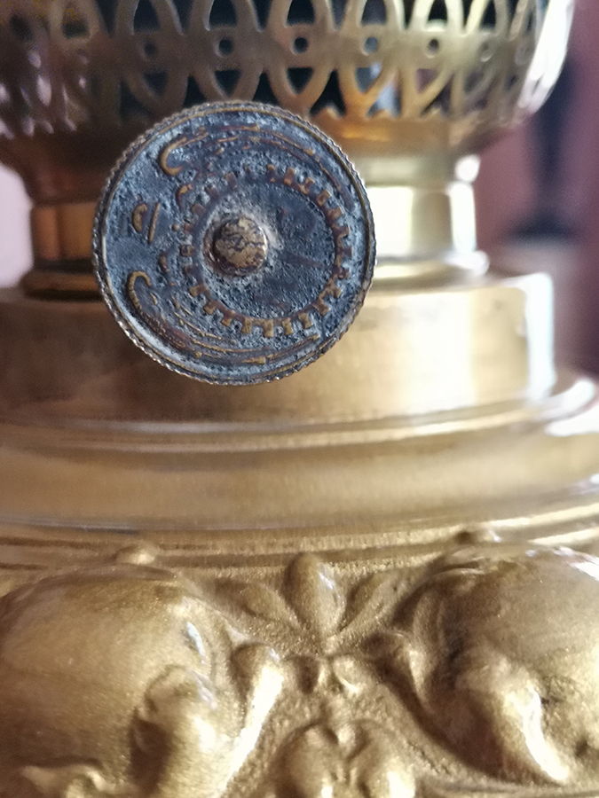 Fotografia kolorowa przedstawia fragment mosiężnego zbiornika lampy naftowej oraz fragment palnika z pinezką pokrętła. Na pokrętle widoczny jest zatarty wzór producenta.