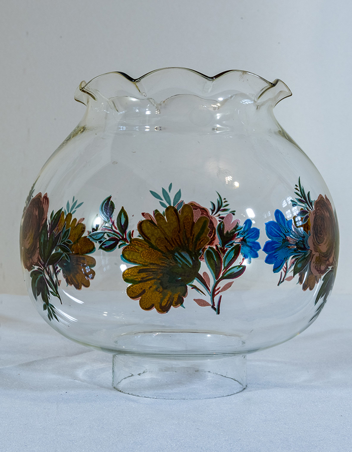 Fotografia kolorowa przedstawia szklany przezroczysty klosz o kwiatowym kształcie, na którym namalowane są kompozycje ze stylizowanych kwiatów w kolorze pomarańczu, zieleni i niebieskim.