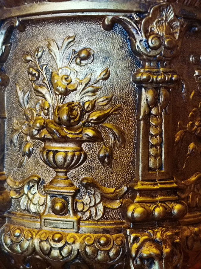 Fotografia kolorowa przedstawia wypukłą dekorację mosiężnego korpusu lampy złożoną z półplastycznych pilastrów, między którymi umieszczone są wazony z bukietami kwiatów.