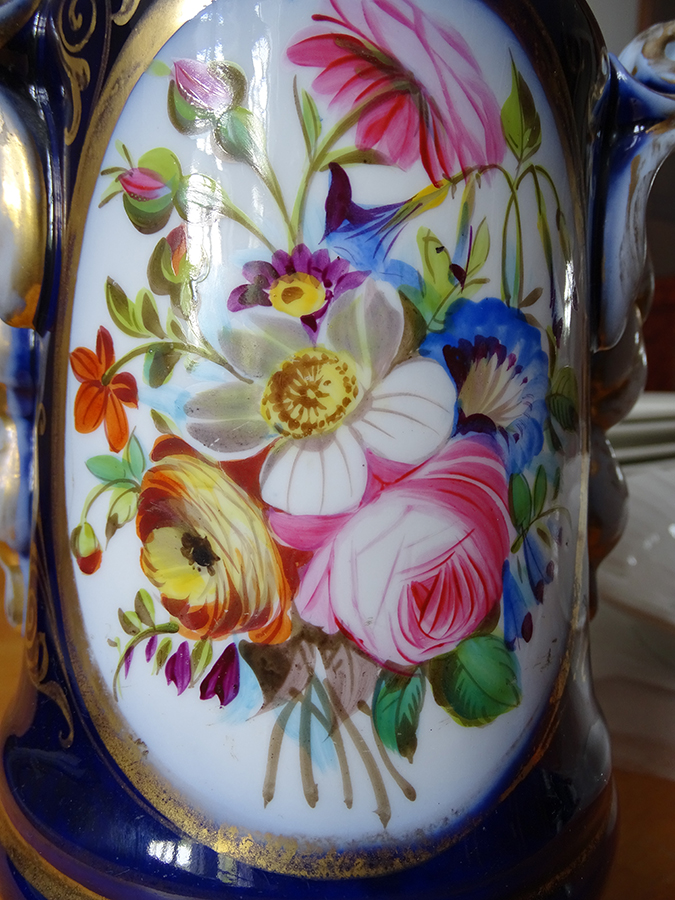 Fotografia kolorowa przedstawia bukiet kwiatowy porcelanowego korpusu lampy naftowej. W owalu bukiet kwiatów w kolorach białym, różowym, niebieskim, pomarańczowym. Pomiędzy kwiatami zielone liście.