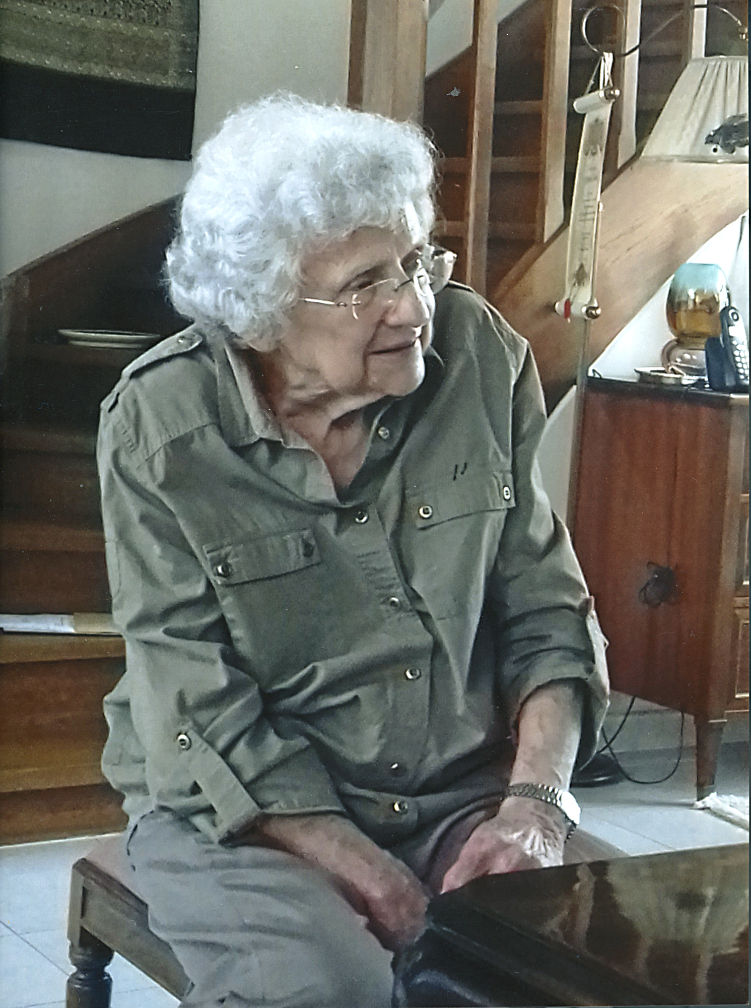 Fotografia współczesna przedstawiająca Janinę Jasińską-Luterek; starsza kobieta, z siwymi, kręconymi włosami do ramion, w okularach, w ujęciu do kolan, siedząca na niskim stołku o rzeźbionych nogach. W tle fragment drewnianych schodów oraz elementy wyposażenia pokoju.