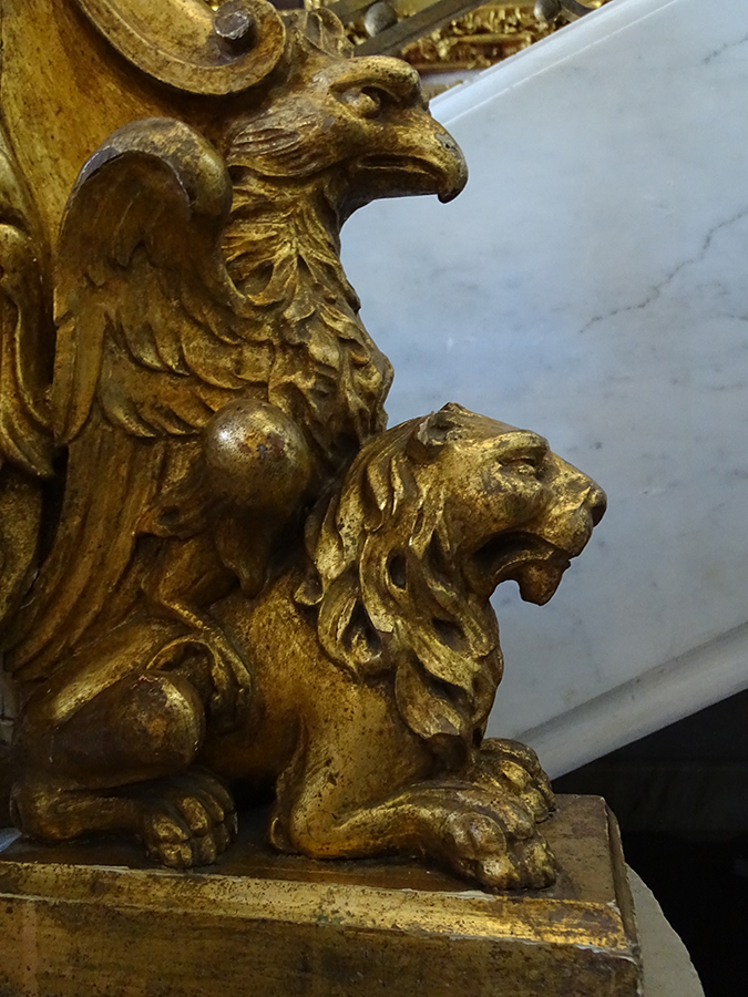Fotografia kolorowa, współczesna przedstawia fragment drewnianego, rzeźbionego złoconego ornamentu w postaci orła orła siedzącego na lwie. Zwierzęta ukazane w profilu. Tło jasne, marmurowe.
