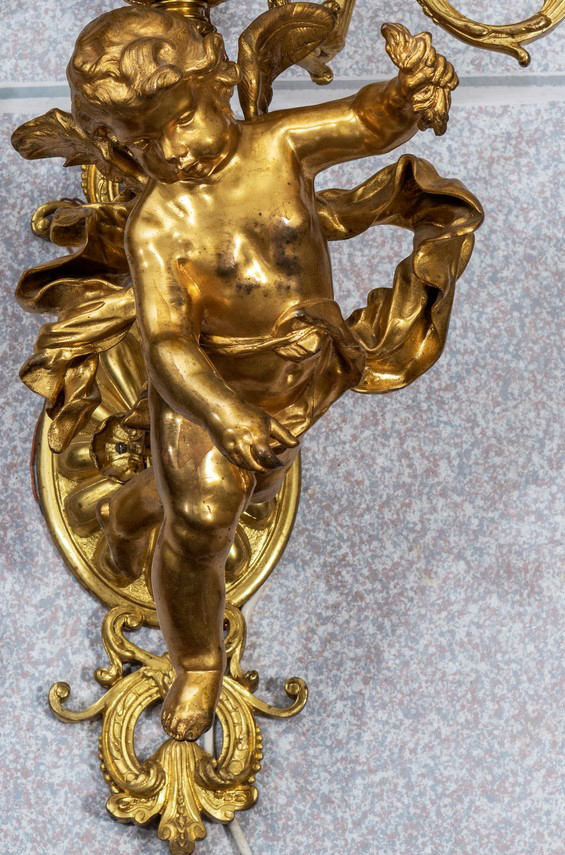 Fotografia kolorowa przedstawia pełnoplastyczną, złoconą figurę amora, na tle szarej ściany.