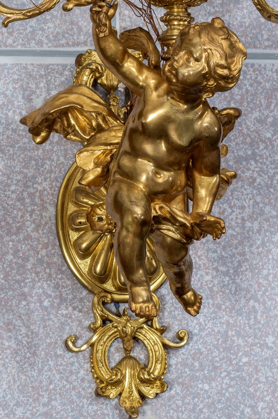 Fotografia przedstawia pełnoplastyczną, złoconą figurę amora, na tle szarej ściany.