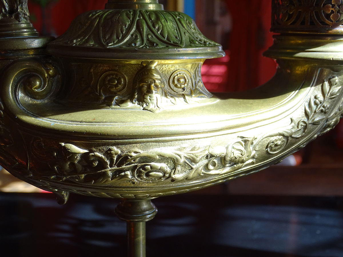 Fotografia kolorowa przedstawia zbliżenie na dekorowany wypukłym ornamentem zbiornik lampy naftowej. Kształt zbiornika niesymetrycznym , łódkowaty.