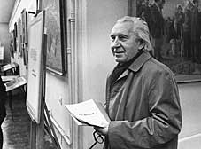 Fotografia czarno-biała przedstawia malarza Bolesława Stawińskiego. Starszy mężczyzna, z dłuższymi siwymi włosami, ukazany jest w półpostaci na tle galerii obrazów. Ubrany jest w płaszcz, w rękach trzyma plik kartek.