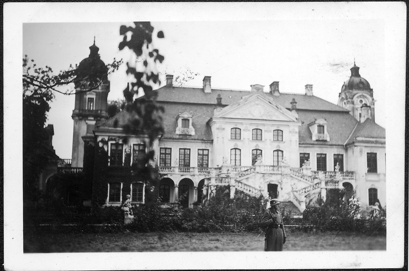 2.	Fotografia archiwalna z lat 40. XX wieku przedstawia pałac w Kozłówce od strony ogrodu. Pałac jednopiętrowy, z wysuniętą częścią środkową , przy której duże schody oraz dwiema wieżami po obu stronach. Na tle pałacu stojący mężczyzna w mundurze żołnierza Wermachtu. Fotografia czarno-biała.