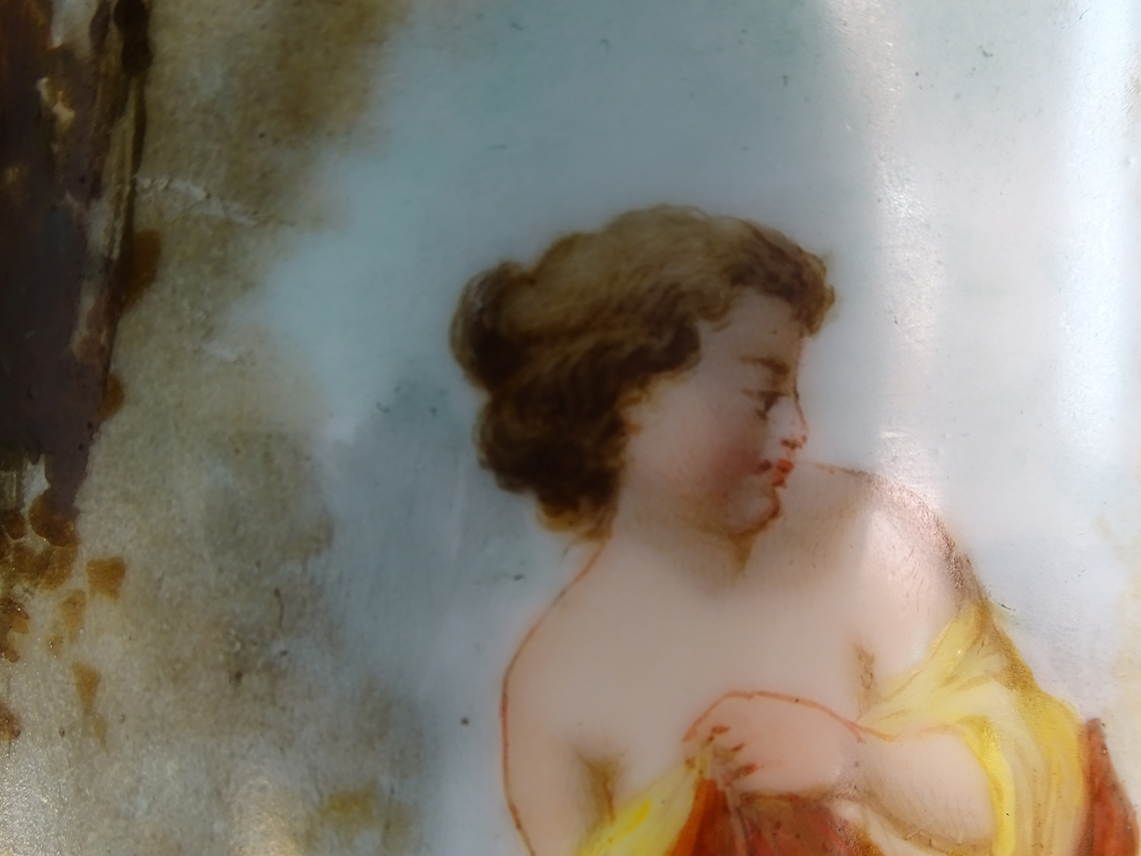 Fotografia kolorowa ukazująca fragment dekoracji malarskiej korpusu lampy naftowej. Przedstawiona jest postać kobieca w ujęciu do ramion, z pomarańczowo-żółtym szalem. Głowa kobiety zwrócona w prawą stronę, jej gęste włosy upięte są w luźny kok. W tle fragmenty leśnego pejzażu.