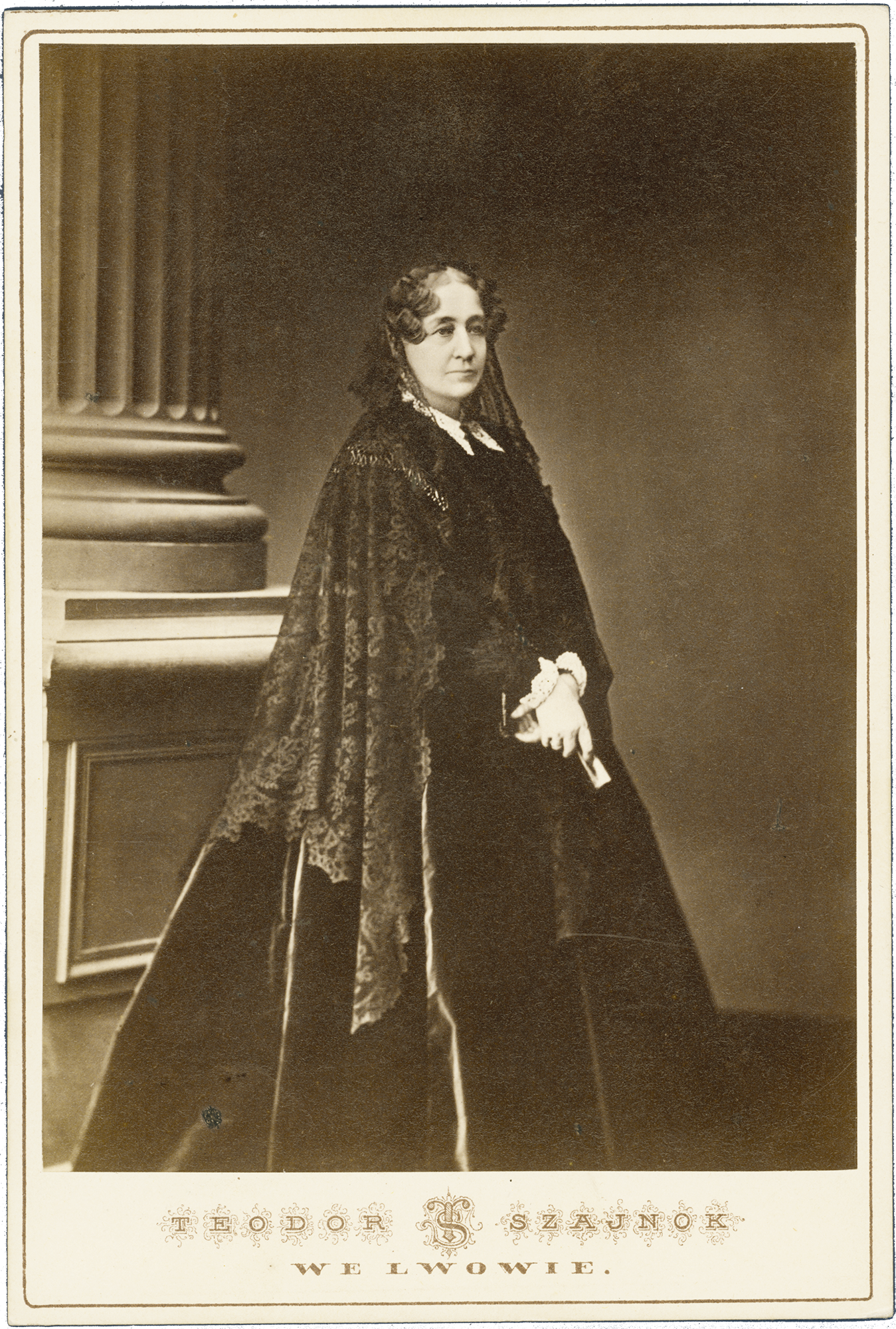 Fotografia archiwalna, czarno biała przedstawiająca dojrzałą kobietę w całej postaci, ubraną w ciemną, długą suknię z czarnym, koronowym welonem na włosach; tło neutralne. Po lewej widoczny fragment kolumny na wysokiej podstawie.