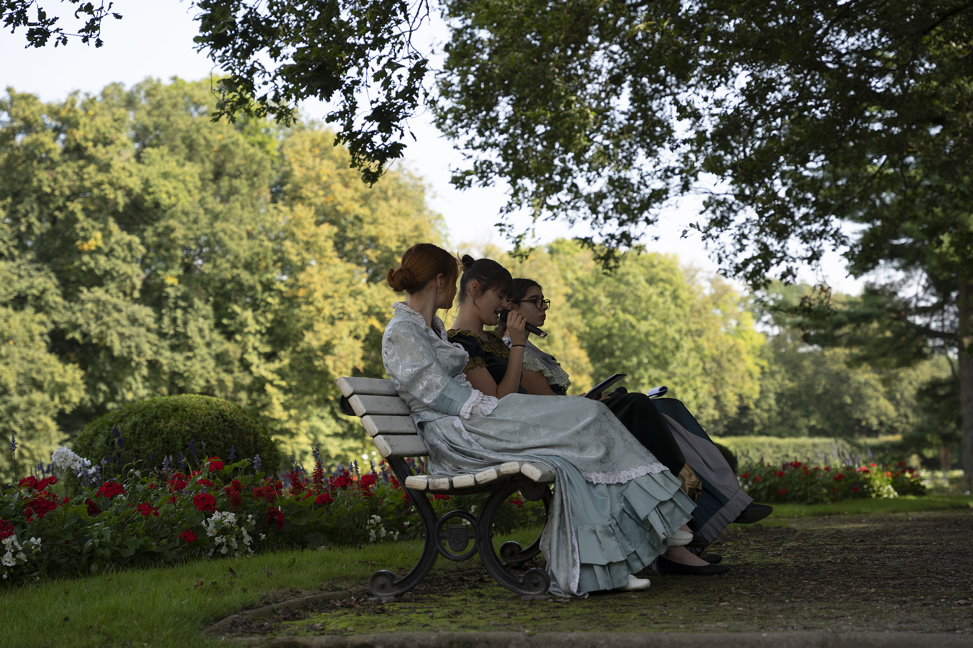 Narodowe Czytanie 2023. Kolorowa fotografia przedstawia trzy dziewczyny w sukniach stylizowanych na koniec XIX wieku. Siedzą na ławce na tle ogrodu, odczytują fragmenty powieści Elizy Orzeszkowej „Nad Niemnem.