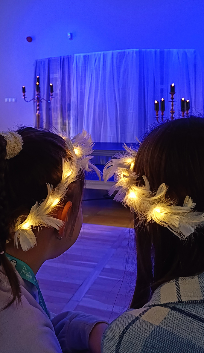 Dwie dziewczynki w ujęciu do ramion, ze świecącymi aureolami na głowach, patrzą w stronę oświetlonej na niebiesko sceny.