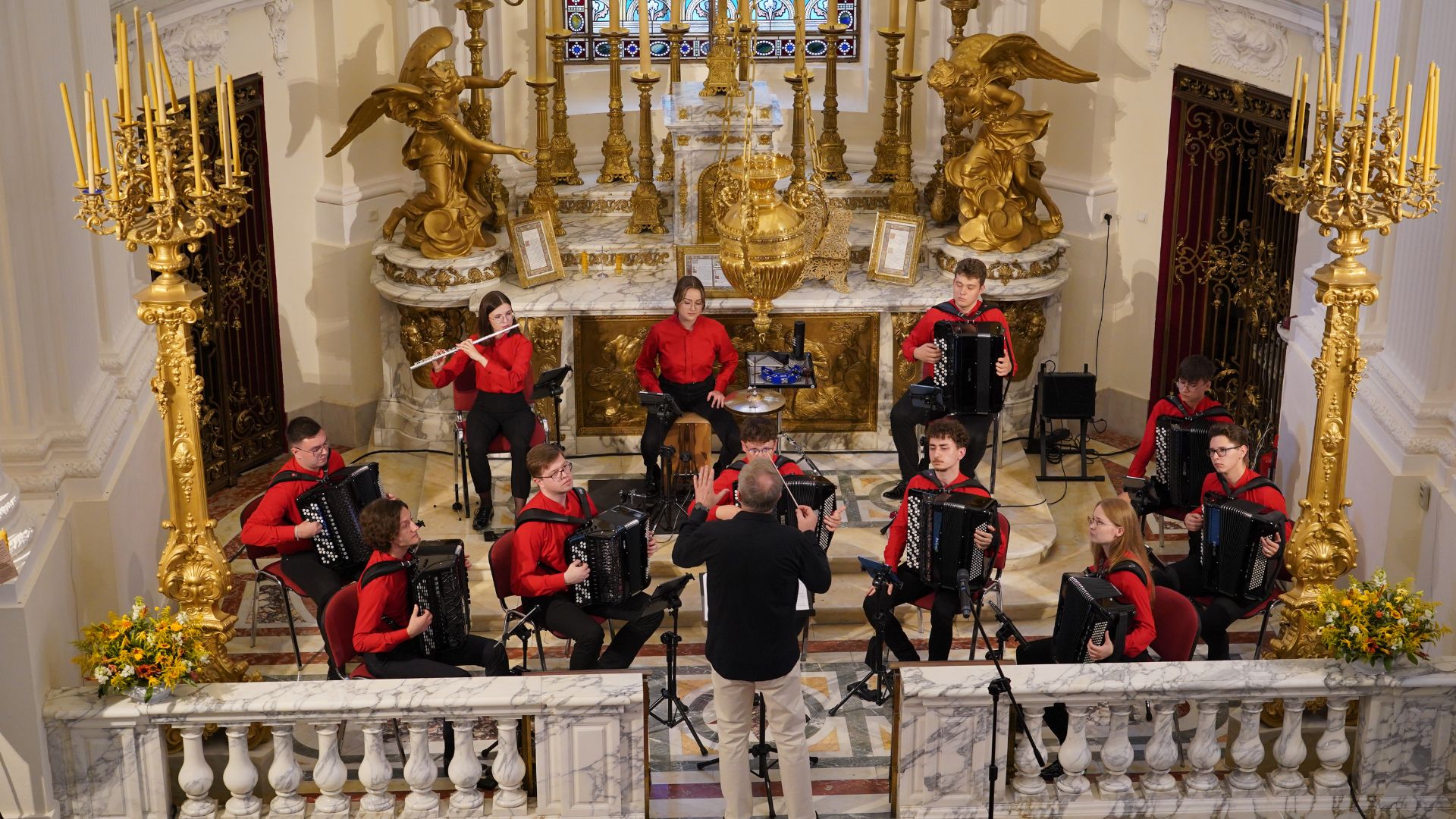 Na zdjęciu widoczny jest występ orkiestry w bogato zdobionym wnętrzu barokowego kościoła. Grupa muzyków, ubrana w czerwono-czarne stroje, gra na akordeonach, flecie i perkusji. Dyrygent, stojący tyłem do widowni, prowadzi zespół, dynamicznie gestykulując.
Tło sceny stanowi złocony ołtarz z barokowymi rzeźbami aniołów, wysokimi świecznikami oraz witrażem, przez który wpada światło. Przed ołtarzem znajduje się marmurowa balustrada, za którą siedzą muzycy. Widownia, ubrana elegancko, słucha koncertu, niektórzy robią zdjęcia.
Cała scena jest dobrze oświetlona, a złote dekoracje nadają miejscu podniosły i uroczysty charakter.