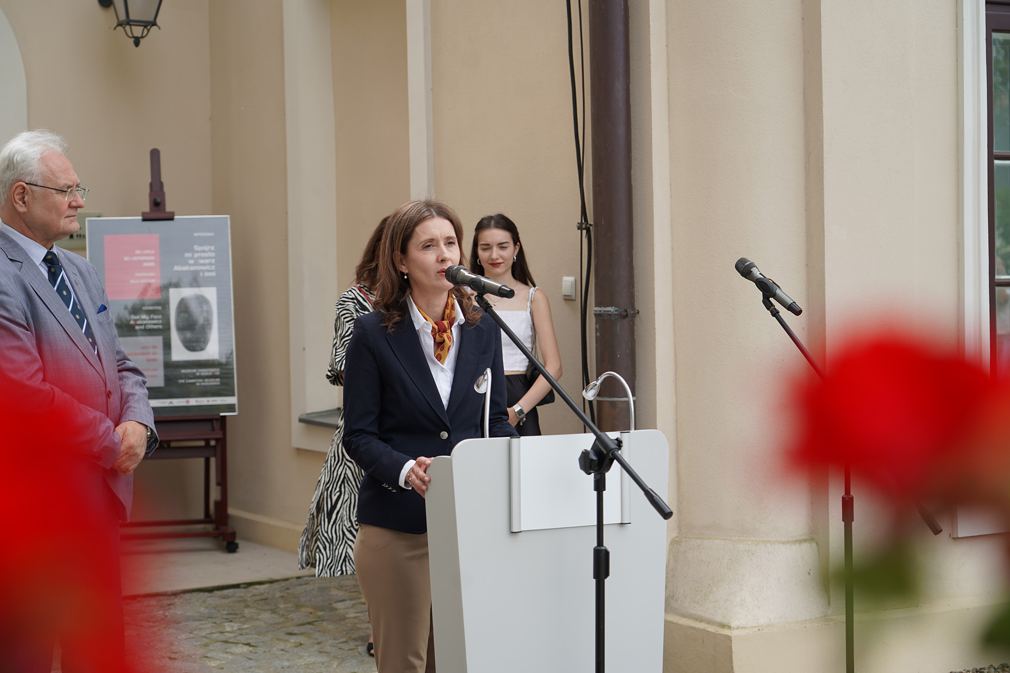 Fot. 2 Fot. 2
Na zdjęciu widzimy scenę z wernisażu, odbywającą się na zewnątrz przy wejściu do eleganckiego, jasnego budynku – prawdopodobnie muzeum lub galerii. Na pierwszym planie, przy białej mównicy z mikrofonem, stoi kobieta w średnim wieku, przemawiająca do zebranych gości. Ubrana jest elegancko: ma na sobie granatową, dopasowaną marynarkę, białą koszulę i ozdobną apaszkę w ciepłych kolorach – brązowo-pomarańczową z żółtymi akcentami. Jej włosy są rozpuszczone, brązowe, lekko falowane. Kobieta patrzy przed siebie z powagą i skupieniem.
Na dalszym planie, po lewej stronie, stoi starszy mężczyzna w szarym garniturze w drobną kratkę, z białymi włosami i okularami – uważnie słucha przemówienia. Za nim znajduje się plakat zapowiadający wystawę „Spójrz mi prosto w twarz. Abakanowicz i inni”, ustawiony na ciemnej sztaludze.
Za przemawiającą kobietą, bliżej ściany budynku, stoją jeszcze dwie osoby: kobieta w czarno-białej sukience w zwierzęcy wzór oraz młoda dziewczyna w białym topie i czarnej, błyszczącej spódnicy, z torebką na ramieniu.
Na dole kadru, lekko rozmyte na czerwono, widoczne są duże kwiaty – prawdopodobnie pelargonie – które wprowadzają do kompozycji kolorystyczny akcent i delikatnie kadrują scenę.
Całość oddaje atmosferę oficjalnego, ale ciepłego wydarzenia plenerowego, związanego z kulturą i sztuką.