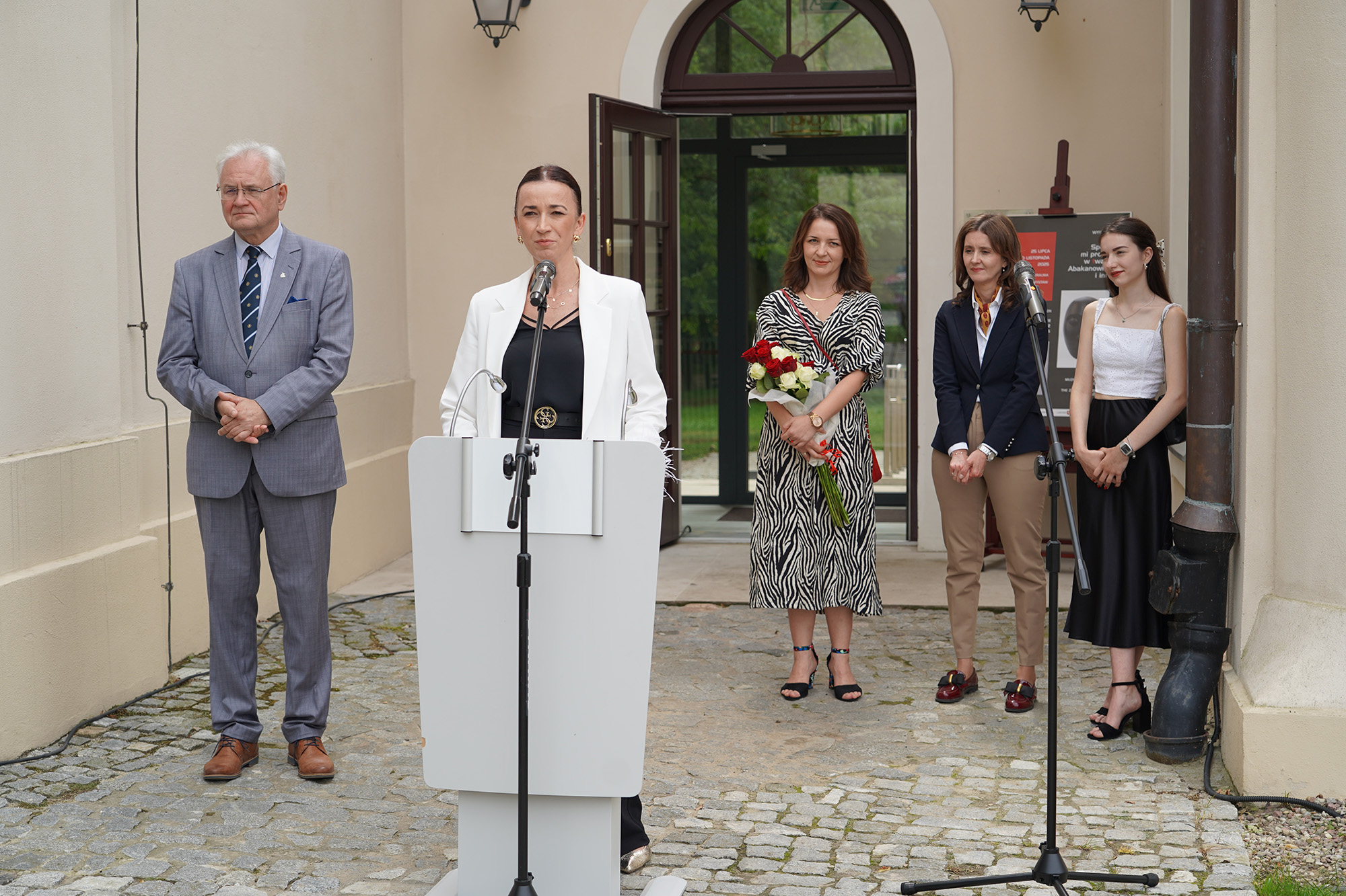 Fot. 4 Fot. 4
Na zdjęciu uchwycony jest moment przemówienia podczas wernisażu wystawy. Scena rozgrywa się na zewnątrz, przed wejściem do eleganckiego budynku z jasną elewacją i przeszklonymi drzwiami.
Na pierwszym planie, na środku zdjęcia, znajduje się kobieta stojąca za białą mównicą i mówiąca do mikrofonu. Ma średniej długości, brązowe włosy i uśmiechniętą twarz. Ubrana jest w sukienkę o wyrazistym, czarno-białym wzorze przypominającym skórę zebry. Przez ramię ma przewieszony czerwony pasek torebki. Kobieta wygląda na pogodną i swobodną, mówi z uśmiechem.
Po lewej stronie zdjęcia, lekko w tle, stoi starszy mężczyzna w jasnoszarym garniturze w subtelną kratkę. Ma siwe włosy, okulary i spokojny wyraz twarzy. Ręce ma splecione przed sobą. Za nim znajdują się otwarte szklane drzwi wejściowe, przez które widać fragment wnętrza – w tym plansze wystawowe z grafikami.
Po prawej stronie zdjęcia, również w tle, stoją dwie kobiety. Jedna z nich ubrana jest w granatową marynarkę, białą koszulę i jasne spodnie. Ma brązowe, falujące włosy i kolorową apaszkę. Obok niej stoi młoda kobieta w białym topie i czarnej, błyszczącej spódnicy. Ma ciemne, długie włosy, lekki makijaż i torebkę na łańcuszku przewieszoną przez ramię. Obie kobiety patrzą z uśmiechem w stronę przemawiającej.
W tle po prawej stronie, przy ścianie, ustawiony jest plakat wystawy zatytułowanej „Spójrz mi prosto w twarz. Abakanowicz i inni”.
Atmosfera zdjęcia jest uroczysta, ale zarazem ciepła i serdeczna – pełna zaangażowania i pozytywnych emocji.