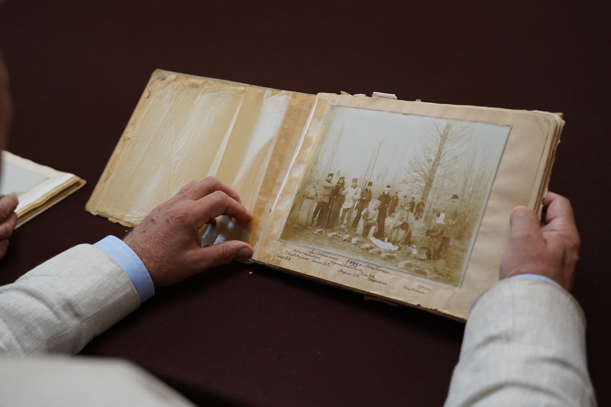 Podpisanie protokołu darowizny przez Pana Andrzeja Belinę Brzozowskiego i Pana Andrzeja Pruszkowskiego, Dyrektora Muzeum Zdjęcie przedstawia podpisanie protokołu darowizny przez Pana Andrzeja Belinę Brzozowskiego i Pana Andrzeja Pruszkowskiego, Dyrektora Muzeum. Na zdjęciu widać dwóch mężczyzn siedzących przy dużym stole pokrytym bordowym obrusem. Mężczyzna po lewej stronie ma białe włosy, okulary i jest ubrany w jasny garnitur z niebieską koszulą i krawatem w ciemne paski. Siedzi prosto, patrząc przed siebie. Mężczyzna po prawej stronie ma siwe włosy, okulary i nosi ciemny polo-shirt. Jest pochylony nad dokumentami, które podpisuje długopisem. Za nimi znajduje się bardzo duży portret w złotej, bogato zdobionej ramie. Obraz przedstawia kobietę w XVIII-wiecznym stroju – ma na sobie bordową suknię z białymi i czarnymi akcentami oraz koronkowy czepek na głowie. Siedzi na ozdobnym fotelu, a obok niej widać królewskie insygnia – koronę oraz berło.