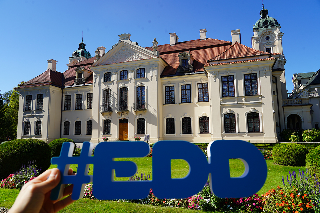 Na zdjęciu widać reprezentacyjny pałac w Kozłówce – siedzibę Muzeum Zamoyskich. Budynek ma jasną, kremową elewację, liczne wysokie okna w ciemnych ramach i czerwony dach kryty dachówką. Zwieńczony jest dekoracyjnymi szczytami oraz wieżyczkami z zielonymi kopułami.
Na pierwszym planie widzimy rękę trzymającą duży, niebieski napis w formie hasztagu: #EDD – nawiązujący do Europejskich Dni Dziedzictwa. W tle, przed pałacem, rozciąga się starannie utrzymany ogród z kolorowymi kwiatami i przyciętymi krzewami.
Całość fotografii, wykonanej w słoneczny dzień, oddaje elegancję rezydencji i podkreśla jej rolę jako miejsca wydarzeń kulturalnych.