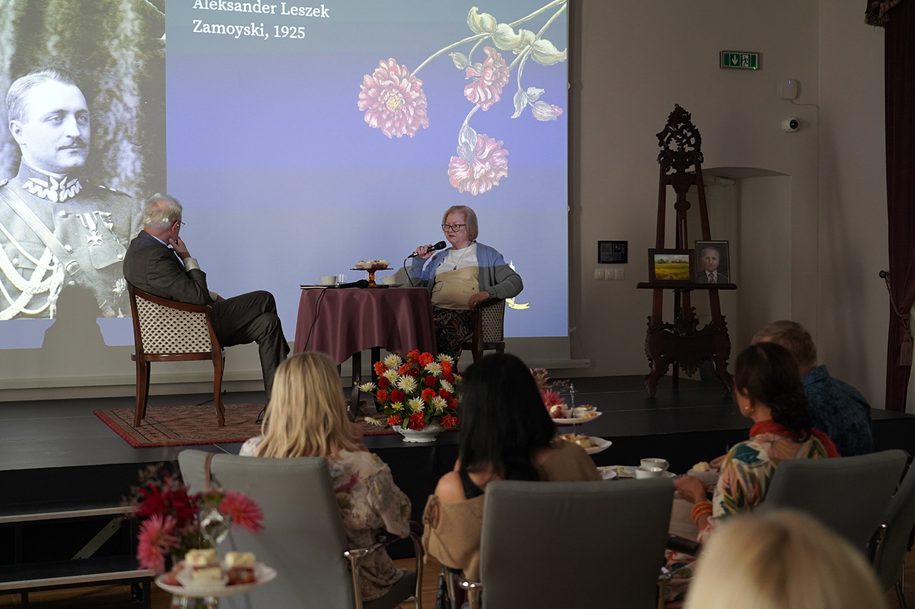 Na fotografii widzimy wydarzenie odbywające się w eleganckiej sali teatralnej Muzeum Zamoyskich w Kozłówce.
Na pierwszym planie, tyłem do obiektywu, siedzą uczestnicy spotkania przy stolikach nakrytych białą zastawą, z filiżankami herbaty oraz paterami z ciastem i kanapkami.
W centrum kadru znajduje się scena. Przy okrągłym stoliku nakrytym bordowym obrusem siedzą dwie osoby. Po lewej stronie, pan Andrzej Pruszkowski, Dyrektor Muzeum Zamoyskich, prowadzący to spotkanie. Mężczyzna siedzi bokiem do publiczności, z uwagą wsłuchując się w opowieść Inki. Po prawej, wspomniana Jadwiga Inka Zamoyska, ubrana w jasny sweter, w okularach, trzyma mikrofon opowiada historię.
Za rozmówcami widoczny jest ekran z dużą projekcją – czarno-białym portretem mężczyzny w mundurze z 1925 roku, podpisanym jako Aleksander Leszek Zamoyski. Jest to ostatni właściciel Kozłówki, tata Inki. Ekran ozdobiony jest także ilustracją kwiatów.
Na scenie, obok ekranu, stoi drewniana sztaluga, na której ustawiono ramki ze zdjęciami – jedno przedstawia portret mężczyzny, drugie pejzaż. Przed stolikiem ustawiono dużą, kolorową kompozycję kwiatową w odcieniach czerwieni, bieli i pomarańczu.