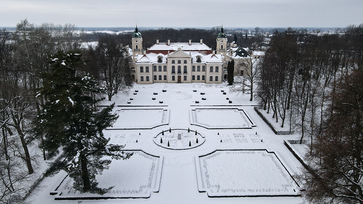 Na fotografii widoczny jest zimowy krajobraz oglądany z lotu ptaka. W centrum kadru stoi duży, symetryczny pałac Zamoyskich w Kozłówce o jasnej, kremowej elewacji. Budynek ma dwie kondygnacje oraz wysokie, ciemne okna rozmieszczone regularnie wzdłuż fasady. Po lewej i prawej stronie pałacu wznoszą się dwie smukłe wieże zwieńczone zielonkawymi kopułami. Dach pałacu pokryty jest warstwą śniegu.
Przed budynkiem rozciąga się rozległy, formalny ogród o geometrycznym układzie. Śnieg dokładnie podkreśla jego kształt. W centrum ogrodu znajduje się okrągły plac z niewielkim pionowym elementem pośrodku, prawdopodobnie pomnikiem lub fontanną. Od tego miejsca odchodzą cztery proste aleje, tworzące kształt krzyża. 
Po obu stronach alei widoczne są prostokątne kwatery ogrodowe, obramowane niskimi, ciemnymi żywopłotami. Roślinność jest zapewne przycięta, lecz obecnie całkowicie przykryta śniegiem. Wzdłuż osi ogrodu ustawione są małe, stożkowate krzewy.
Cały ogród otaczają wysokie, bezlistne drzewa, tworzące park. W tle, za pałacem, rozciąga się płaski, zimowy krajobraz z polami i zabudowaniami ledwo widocznymi w oddali. Niebo jest jasnoszare i pochmurne. Obraz sprawia wrażenie spokojnego, chłodnego i bardzo uporządkowanego.