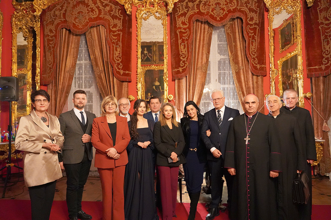 Zdjęcie przedstawia grupowy portret wykonany w reprezentacyjnej, pałacowej sali o bogatym wystroju. Tło stanowią czerwone ściany, wysokie okna z białymi firanami oraz ciężkie, bordowe zasłony z dekoracyjnymi lambrekinami. Pomiędzy oknami widoczne są duże, pionowe lustra w złoconych, rzeźbionych ramach oraz obrazy w ozdobnych ramach. Wnętrze jest symetryczne i silnie dekoracyjne, o ciepłej, złoto-czerwonej kolorystyce.
Na pierwszym planie, na czerwonym dywanie, stoi grupa kilkunastu osób ustawionych w jednym rzędzie, blisko siebie, zwróconych twarzami do obiektywu. Są to kobiety i mężczyźni w różnym wieku, ubrani elegancko i formalnie. Kobiety mają na sobie sukienki, garnitury lub eleganckie komplety w stonowanych kolorach: beżu, czerwieni, granatu i czerni. Mężczyźni ubrani są w garnitury, marynarki lub ciemne stroje duchowne.
Po prawej stronie grupy stoją duchowni w czarnych sutannach; jeden z nich ma na piersi widoczny krzyż na łańcuchu. Pozostałe osoby ubrane są świecko, w stylu oficjalnym, odpowiednim do uroczystego wydarzenia. Większość osób ma neutralny lub lekko uśmiechnięty wyraz twarzy i spokojną, wyprostowaną postawę, z rękami swobodnie opuszczonymi lub splecionymi przed sobą.
Całość sprawia wrażenie pamiątkowego zdjęcia wykonanego po oficjalnym wydarzeniu kulturalnym lub uroczystości, w eleganckiej, historycznej przestrzeni muzealnej lub pałacowej.