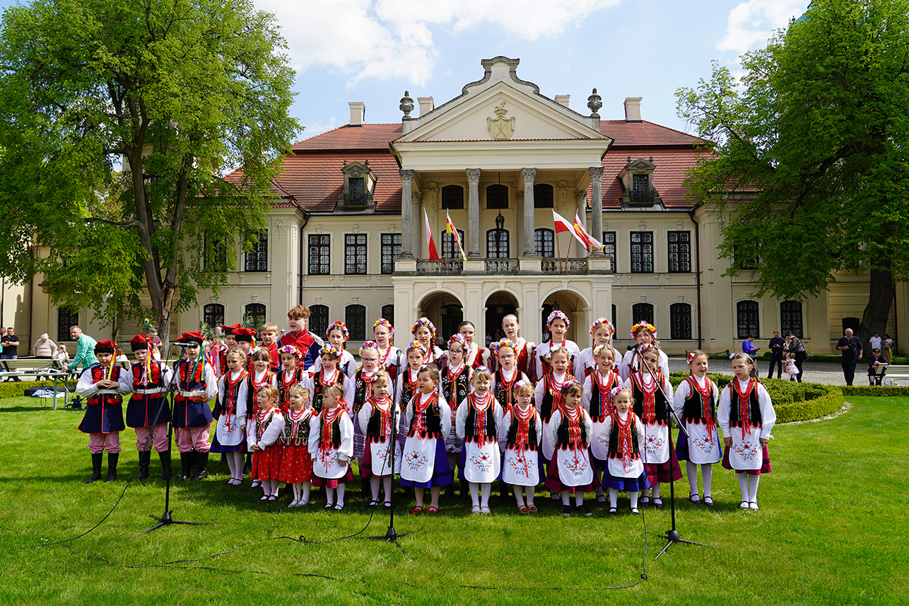 Grupa dzieci w strojach ludowych stoi przed pałacem. Przed nimi, na trawie znajdują się mikrofony.